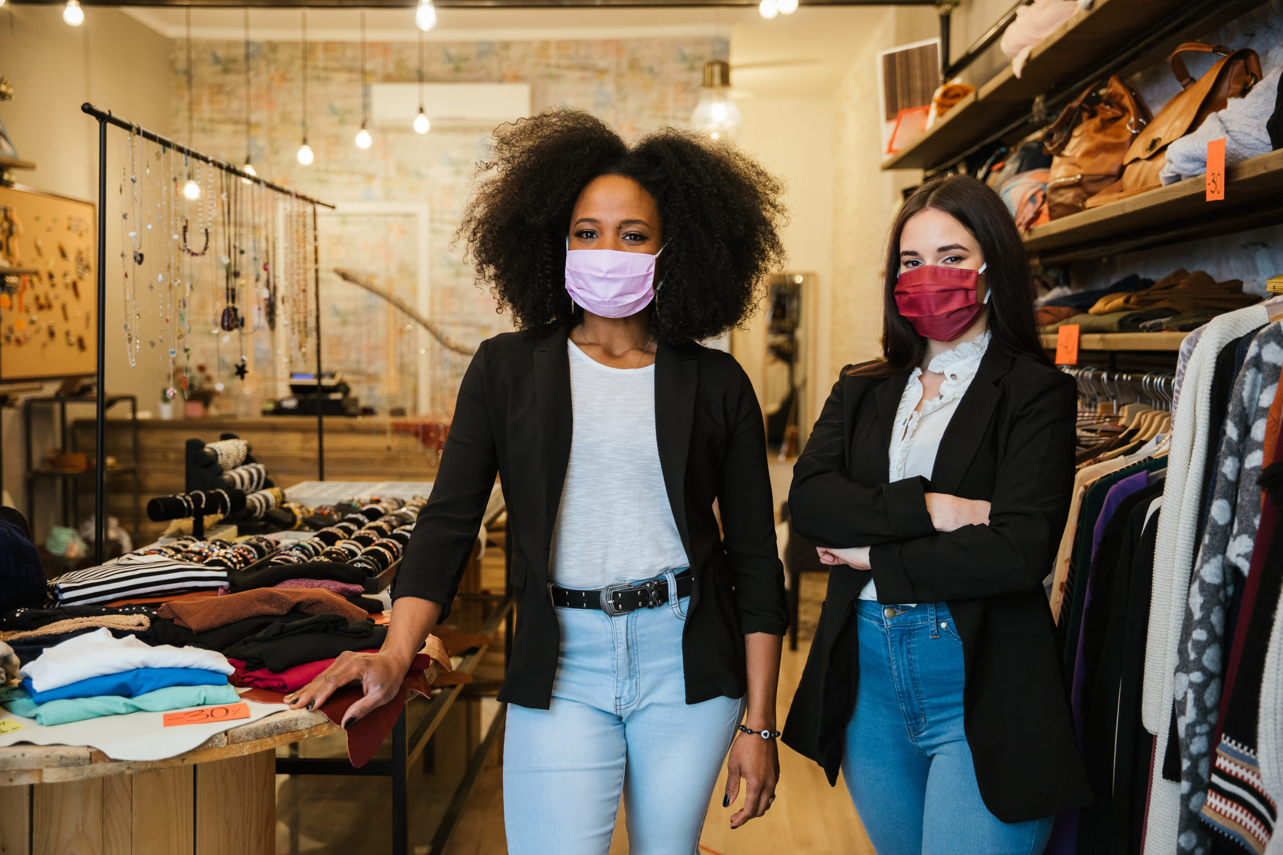 Portrait of two women owners of the clothes shop at the entrance to welcome customers during the Coronavirus Covid 19 pandemic wearing protective face masks – Millennial initiate a start up business